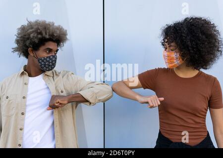 Anonymer junger afroamerikanischer Mann und Frau mit lockigen Haaren in legerer Kleidung und Schutzmasken, die die Ellbogen an der weißen Wand berühren Stockfoto