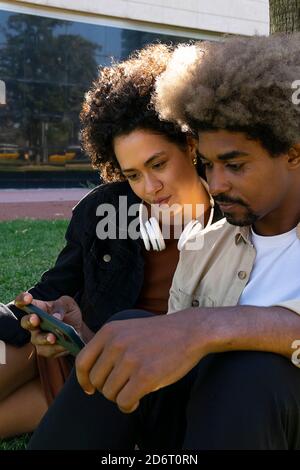 Positive junge afroamerikanische Millennials in trendigen Outfits beim Video Auf dem Smartphone, während Sie im Park auf dem Rasen sitzen Stockfoto