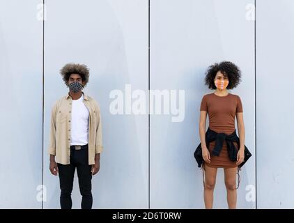 Ruhige junge afroamerikanische Dame und Kerl mit lockigen Haaren In lässiger Kleidung und Gesichtsmasken Blick auf die Kamera während In der Nähe einer schäbigen Betonwand stehen o Stockfoto