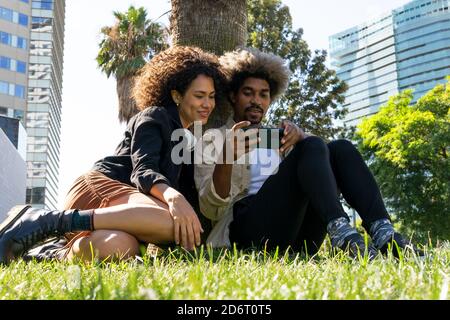 Niedriger Winkel der positiven jungen afroamerikanischen Millennials in trendy Outfits ansehen Video auf dem Smartphone, während auf Gras sitzen in parken Stockfoto