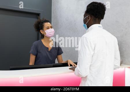 Rückansicht des schwarzen männlichen Patienten in der Maske, die an steht Rezeption in der Nähe von Ärztin vor der medizinischen Behandlung in der Klinik Stockfoto