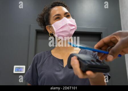 Niedriger Winkel der afroamerikanischen weiblichen Manager in medizinische Maske Empfang der Zahlung über POS-Terminal und Mobiltelefon kontaktlose Zahlung Von Ernte männlichen pa Stockfoto