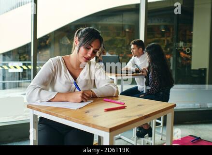 Junge Frau, die mit Stift auf Papier schreibt, während sie an einem Projekt am Tisch im Gebäude arbeitet Stockfoto