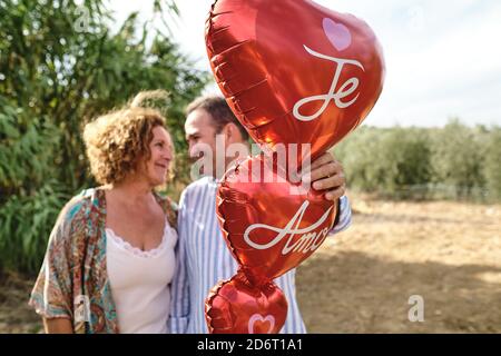 Zartes reifes Paar, das zusammen im Garten mit roter Luft steht Luftballons in Form von Herz und Blick auf einander Während des Spaziergangs Stockfoto