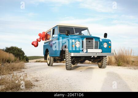 Niedriger Winkel von Vintage glänzendes Auto mit roten Luftballons In Form von Herz geparkt auf Sandstraße in der Landschaft Stockfoto