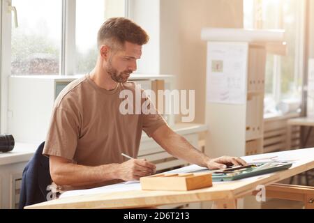 Porträt eines reifen bärtigen Architekten, der Blaupausen ansieht, während er im Sonnenlicht am Zeichenschreibtisch sitzt und Raum kopiert Stockfoto