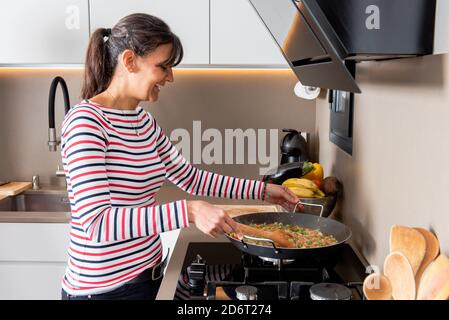 Seitenansicht lachende Frau in legerer Kleidung, die gemischtes Gemüse in der Bratpfanne rührt, während sie das Abendessen in einer stilvollen Küche kocht Stockfoto