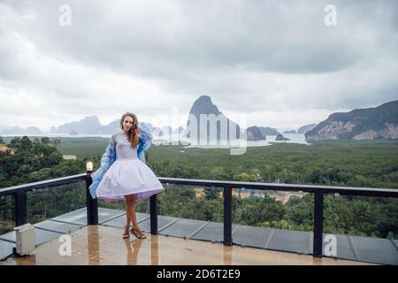 Thailand - 2019. Standpunkt von Samet nangshe. Frau steht auf dem Platz in einem fliegenden Kleid und einem Regenmantel. Das Wetter ist trüb. Stockfoto