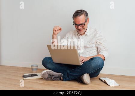 Ganzkörper entzückt reifen Mann tragen lässige Kleidung sitzt auf Boden mit zeitgemäßem Laptop auf Runden und Triumphgesten Stockfoto