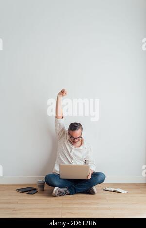 Ganzkörper entzückt reifen Mann tragen lässige Kleidung sitzt auf Boden mit modernem Laptop auf dem Schoß und Ausstrecken der Arme Triumph Stockfoto