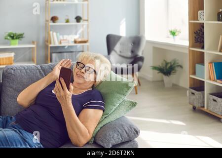 Glückliche lächelnde ältere Frau, die auf dem Sofa lag und sich unterhielt Videoanruf über Telefon mit Familie Stockfoto