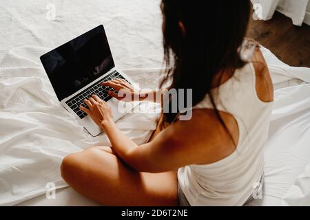 Von oben Rückansicht von unkenntlich Weibchen in Schlafbekleidung sitzend Im Bett und im Laptop nach dem Aufwachen im hellen Schlafzimmer Morgens Stockfoto