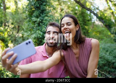 Junge Frau und bärtiger Kerl ragen aus der Zunge, während sie Selfie auf dem Mobiltelefon während des Dates im grünen Park nahmen Stockfoto