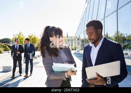 Fokussierte junge Managerin in Anzug zeigt Dokumente an Afrikaner Amerikanischer männlicher Kollege, der in der Nähe eines modernen Gebäudes mit Laptop steht Sonniger Tag Stockfoto