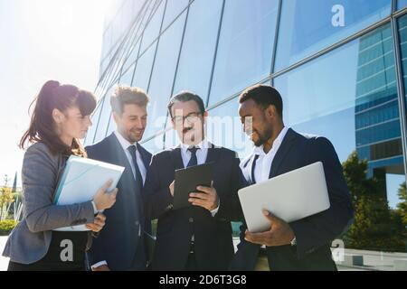 Gruppe verschiedener Kollegen in formellen Anzügen, die Daten zu analysieren Tablet, während Sie zusammen auf der Straße in der Nähe eines modernen Gebäudes mit stehen Verschiedene Gadgets und d Stockfoto
