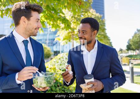 Positive junge, vielfältige Mitarbeiter in formeller Kleidung lächeln beim Haben Mittagspause zusammen auf der Straße an sonnigen Tag Stockfoto