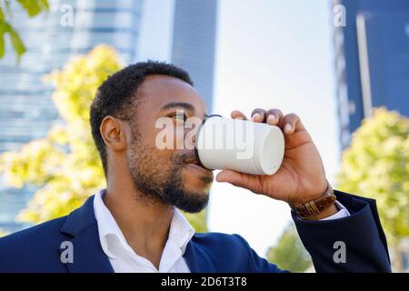 Niedriger Winkel der positiven jungen afroamerikanischen männlichen Manager in Formelles Outfit, das während der Pause Kaffee zum Mitnehmen auf der Straße trinkt Sonniger Tag Stockfoto