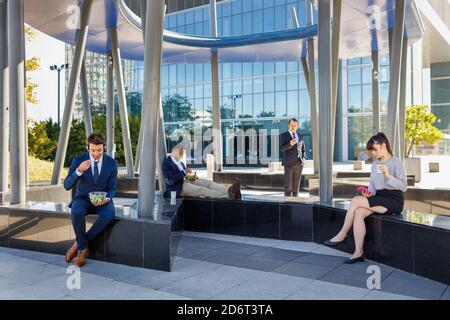 Gruppe von jungen multirassischen Kollegen in stilvollen Outfits sitzen auf Bank in der Nähe des modernen Geschäftszentrums und Mittagessen Stockfoto