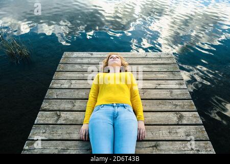Hoher Winkel der ruhigen Frau in Freizeitkleidung liegend auf Hölzerner Pier in der Nähe des Sees und genießen Sie die Stille am Sommertag Stockfoto