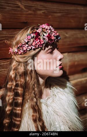 Charming zuversichtlich nordischen jungen Weibchen mit langen welligen blonden Haaren Tragen weißen Pelzmantel und bunten Blumenkranz stehen dagegen Holzwand und lo Stockfoto