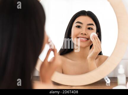 Lächelnde asiatische Frau, die ihr Gesicht putzt, mit Wattepads Stockfoto