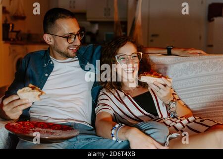 Vorderansicht von jungen multirassischen Paar in lässiger Kleidung Kuscheln Und Pizza essen, während man zusammen auf der Couch sitzt und zuschaut Film auf einem Laptop Stockfoto