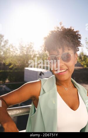 Fröhliche junge afroamerikanische Frau mit Afro-Frisur und trendy Sonnenbrillen, die sich am Geländer am Ellenbogen lehnen und dabei auf die Kamera blicken Genießen Sie den sonnigen Sommer Stockfoto