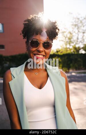 Fröhliche junge afroamerikanische Frau mit Afro-Frisur und trendy Sonnenbrillen, die sich am Geländer am Ellenbogen lehnen und dabei auf die Kamera blicken Genießen Sie den sonnigen Sommer Stockfoto