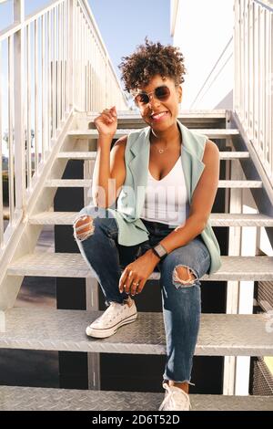Niedriger Winkel der fröhlichen jungen afroamerikanischen Hipster weiblich in Trendy Outfit und Sonnenbrille sitzen auf Treppen auf der städtischen Straße Stockfoto