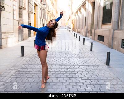 Seitenansicht einer jungen, sportlichen, barfüßigen ethnischen Frau, die lächelt, während sie auf der Straße zwischen alten Gebäuden im Sonnenlicht tanzt Stockfoto