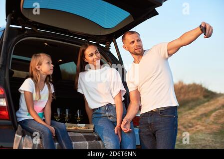 Vater nimmt Selfie seiner schönen Familie im Kofferraum sitzen und mit Pizza und Wein, Familie aussehen weißen T-Shirts und blauen Jeans Stockfoto