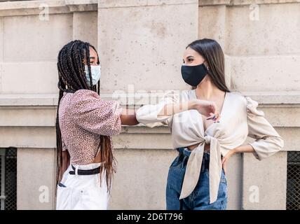 Seitenansicht von positiven jungen multirassischen weiblichen Freundinnen in trendy Kleidung und Schutzmasken begrüßen sich gegenseitig mit Ellenbogenstoß An der City Street Durin Stockfoto