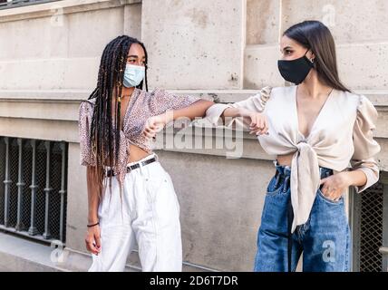 Seitenansicht von positiven jungen multirassischen weiblichen Freundinnen in trendy Kleidung und Schutzmasken begrüßen sich gegenseitig mit Ellenbogenstoß An der City Street Durin Stockfoto