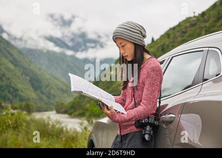 Seitenansicht der jungen nachdenklichen asiatischen Frau in Sportswear ruhen Auf dem Auto mit Buch im grünen bewölkten Tal Stockfoto