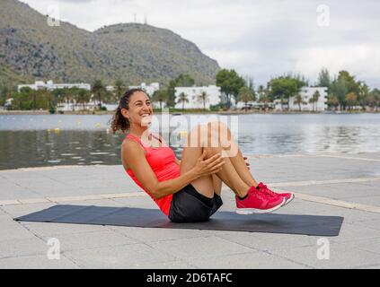 Fröhliche Sportlerin in Sportkleidung, die auf der Matte am Ufer liegt Und dabei Bauch Crunches während des aktiven Trainings im Sommer während Wegschauen Stockfoto