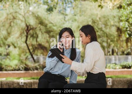 Seitenansicht einer fokussierten asiatischen Geschäftsfrau, die im grünen Park Informationen auf dem Tablet einer glücklichen Kollegin zeigt, die auf dem Smartphone spricht Stockfoto