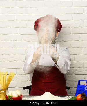 Kochen Sie mit versteckten Gesicht in burgunderroten Uniform sitzt am Tisch mit Pasta und Mehl. Der Koch bereitet Teig zu. Konzept des Kochvorgangs. Mann mit Bart wirft Mehl bis machen Wolke auf weißen Backstein Wand Hintergrund. Stockfoto