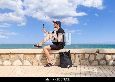 Seitenansicht des fröhlichen Mannes in schwarz tragen sitzen auf Zaun der Böschung in der Nähe des Meeres, während mit Videoanruf Handy unter blauem bewölktem Himmel Stockfoto