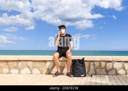 Männchen in schwarz tragen sitzen auf Zaun der Böschung in der Nähe Meer, während Telefonat auf Handy unter blau bewölkt Himmel Stockfoto