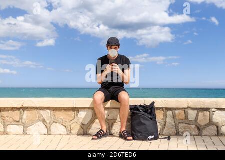 Männchen in schwarz tragen sitzen auf Zaun der Böschung in der Nähe Meer während SMS auf Handy unter blau bewölktem Himmel Stockfoto