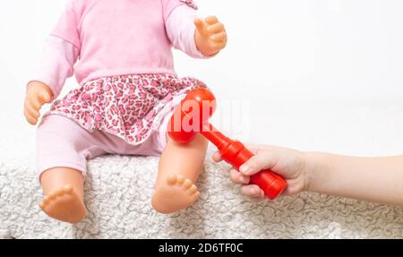 Ein kleines Mädchen spielt mit einer Puppe, und mit einem kleinen neurologischen Hammer trifft es auf das Knie der Puppe. Konzept der Kinderneurologie, Reflexkontrolle Stockfoto