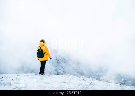 Rückansicht von anonymen aktiven Reisenden in hellgelb warm Jacke mit Rucksack hält Trekkingstöcke und medizinische Maske während Stehen auf verschneiten hil Stockfoto