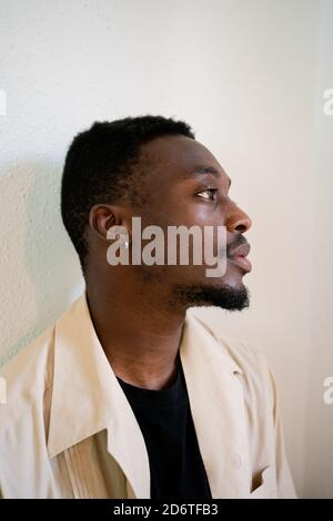 Nachdenkliche junge afroamerikanische männlich in hellbeige formelles Hemd Rücksichtsvoll wegschauen, während man in der Nähe einer weißen Wand steht Stockfoto