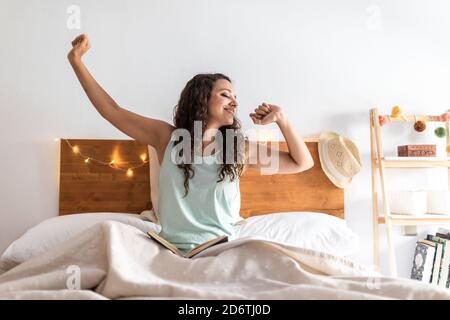 Lächelnde Frau in Schlafkleidung sitzt auf dem Bett unter Decke und Nach dem Erwachen mit geschlossenen Augen sanft dehnen Stockfoto