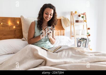 Hoher Winkel der heiteren Frau in Schlafanzug auf dem Bett sitzen Mit einer Tasse frischen Kaffee und Buch lesen beim Genießen Ruhiges Wochenende am Morgen Stockfoto