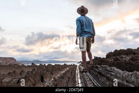 Rückansicht des anonymen männlichen Reisenden, der auf felsiger Küste läuft Nahe welliges Meer unter hellem bewölktem Himmel bei Sonnenuntergang Stockfoto