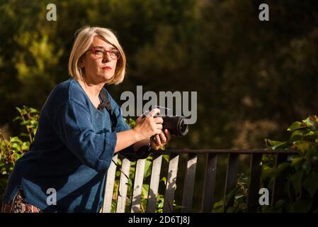 Seitenansicht der seriösen erwachsenen blonde weibliche Fotograf mit professionellen Fotokamera, die sich am Zaun lehnt und im Stehen wegschaut Gegen grünen Bus Stockfoto