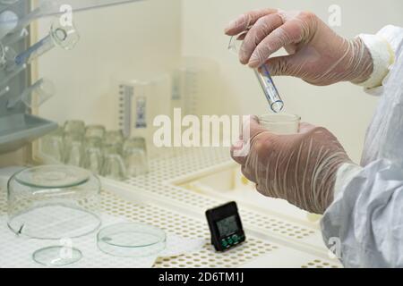 Wissenschaftler mit weißem Schutzmantel, Maske und Handschuhen analysiert eine Virus- oder Bakterienprobe in einem Labor mit Fläschchen, Glasflaschen und Chemikalien Stockfoto