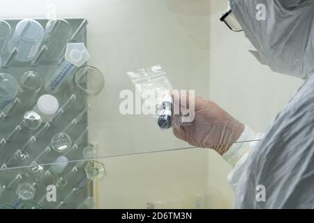 Wissenschaftler mit weißem Schutzmantel, Maske und Handschuhen analysiert eine Virus- oder Bakterienprobe in einem Labor mit Fläschchen, Glasflaschen und Chemikalien Stockfoto