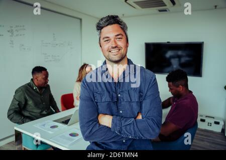 Kaukasischer Geschäftsmann, der stolz mit den Kreuzarmen davor steht Kollegen im Vorstandszimmer während der Teambesprechung Stockfoto